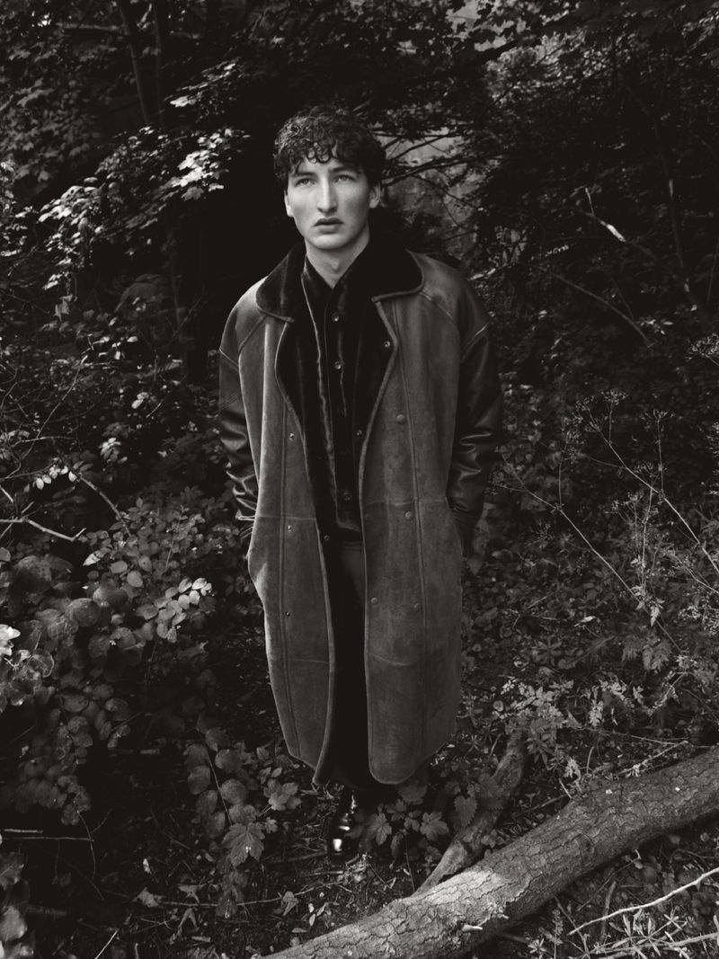 Young man in a long dark shearling coat standing amidst dense woodland foliage in a monochrome photograph.