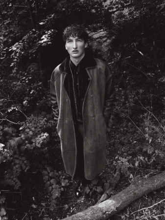 Young man in a long dark shearling coat standing amidst dense woodland foliage in a monochrome photograph.