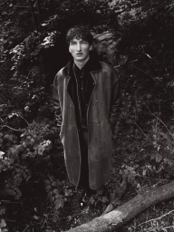 Young man in a long dark shearling coat standing amidst dense woodland foliage in a monochrome photograph.