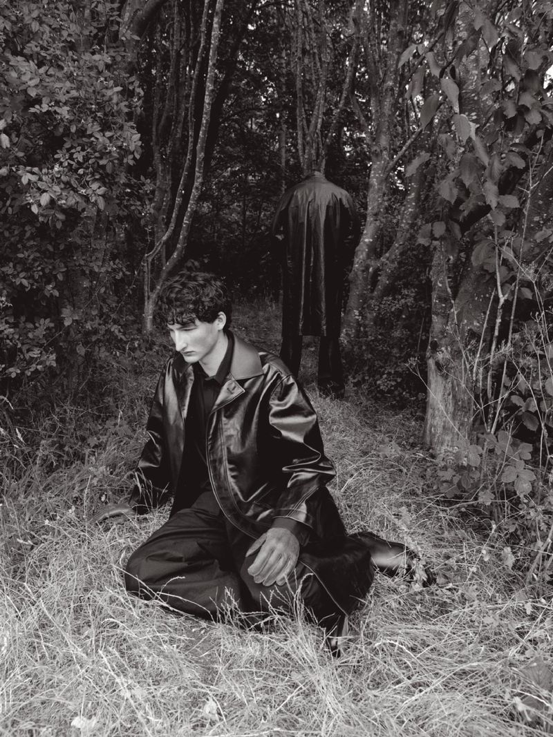 Man kneeling in a dark coat amongst woodland grass with a second figure standing in the background.