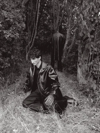 Man kneeling in a dark coat amongst woodland grass with a second figure standing in the background.