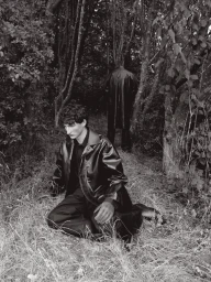 Man kneeling in a dark coat amongst woodland grass with a second figure standing in the background.