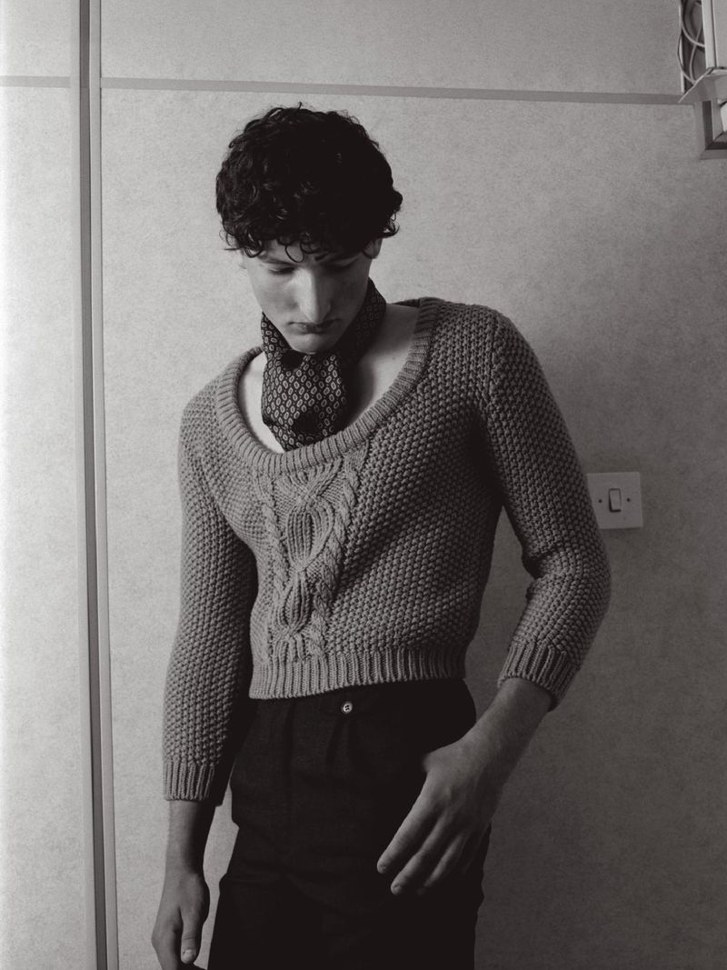 Young man with curly dark hair wearing a patterned scarf and textured sweater standing against a plain interior wall