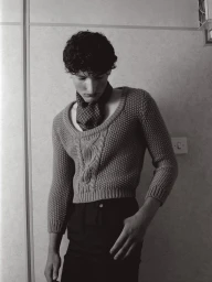 Young man with curly dark hair wearing a patterned scarf and textured sweater standing against a plain interior wall
