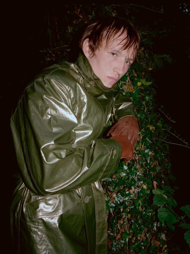 Man wearing a glossy green coat and brown leather gloves posing against dense dark foliage at night.