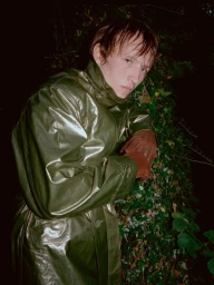 Man wearing a glossy green coat and brown leather gloves posing against dense dark foliage at night.