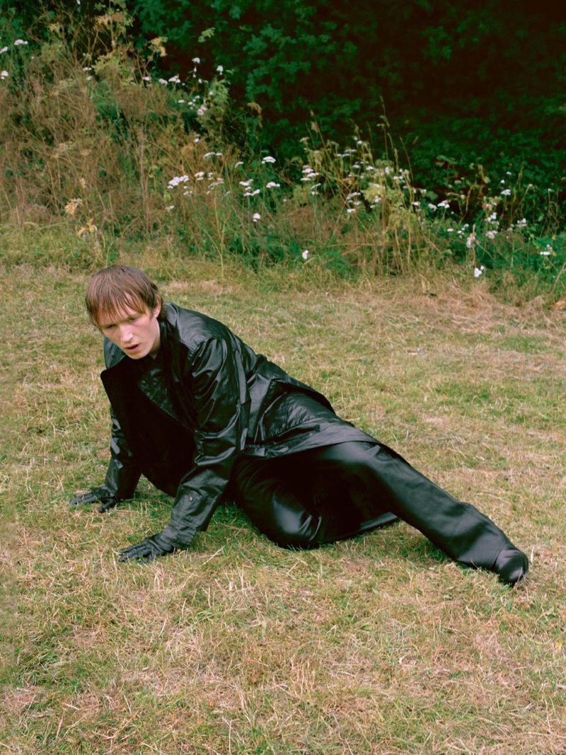 Young man in a black leather coat kneeling on a grassy patch with tall weeds and wildflowers in the background.