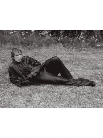 Man in dark jacket and trousers lying on grassy ground outdoors in a black and white editorial photograph.