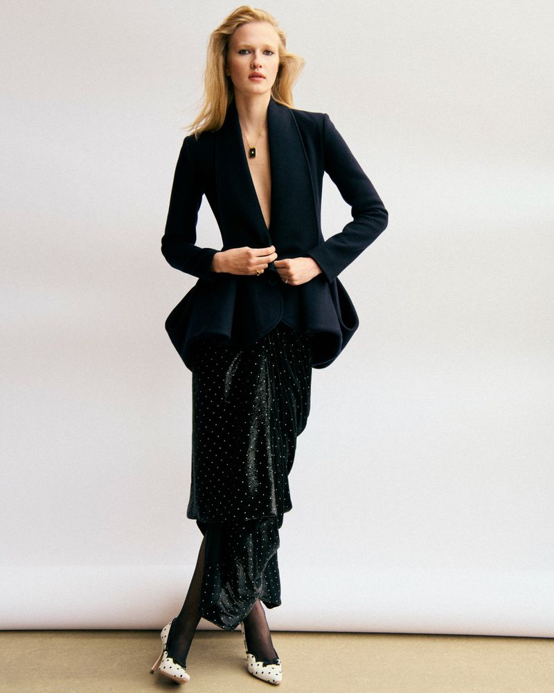 Model wearing a dark blazer and patterned skirt posing against a white seamless background for Elle Sweden