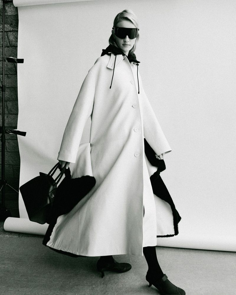 Model in oversized coat and dark sunglasses holding a black handbag against a white seamless backdrop