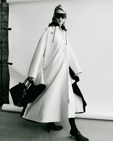 Model in oversized coat and dark sunglasses holding a black handbag against a white seamless backdrop