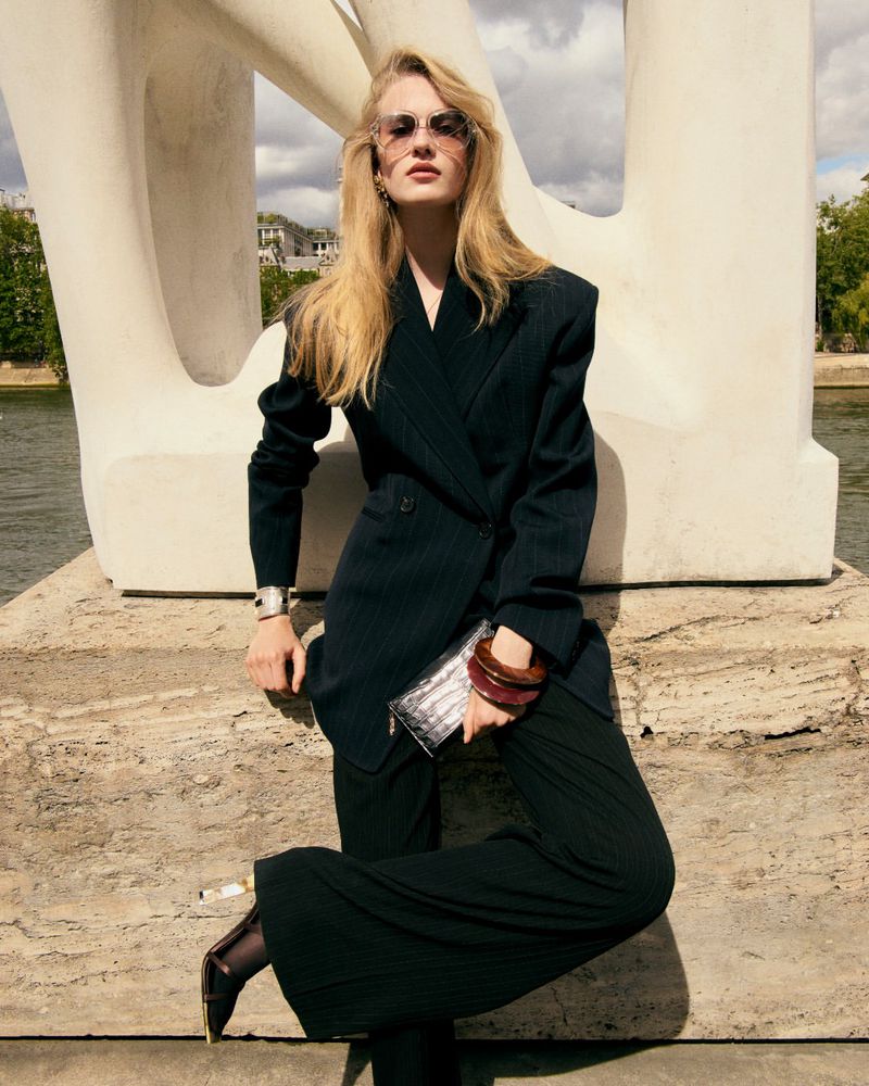 Model in dark pinstripe suit wearing sunglasses and holding a clutch against a large white sculptural monument outdoors.