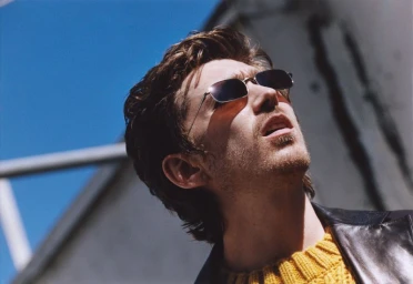 Man wearing sunglasses and yellow sweater looking up against a bright blue sky and structural background.