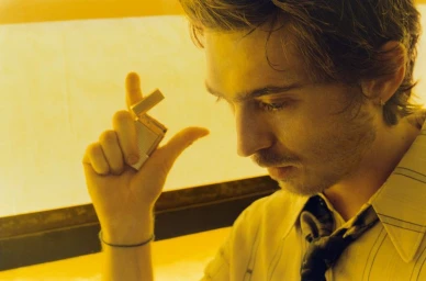 Close-up portrait of Austin Abrams holding a small lighter against a warm yellow-lit background for British GQ.