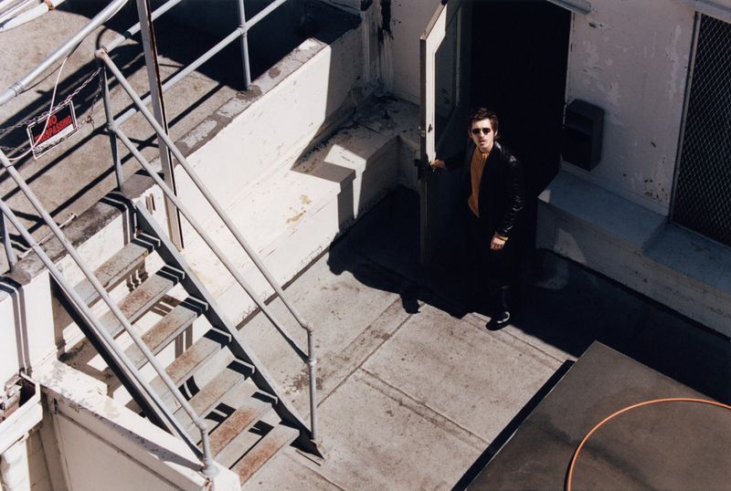 Austin Abrams holding an open doorway on a concrete rooftop with exterior metal stairs, captured from a high angle by photographer Lindsay Ellary.