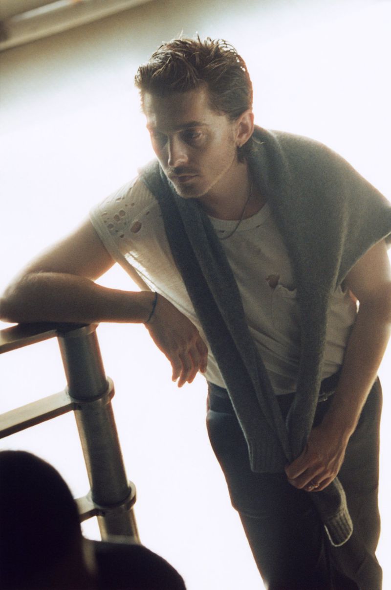 Portrait of Austin Abrams leaning on a railing wearing a white t-shirt and a grey sweater draped over his shoulders for British GQ