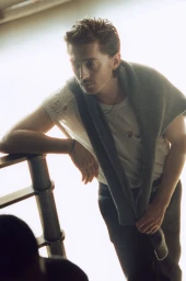 Portrait of Austin Abrams leaning on a railing wearing a white t-shirt and a grey sweater draped over his shoulders for British GQ