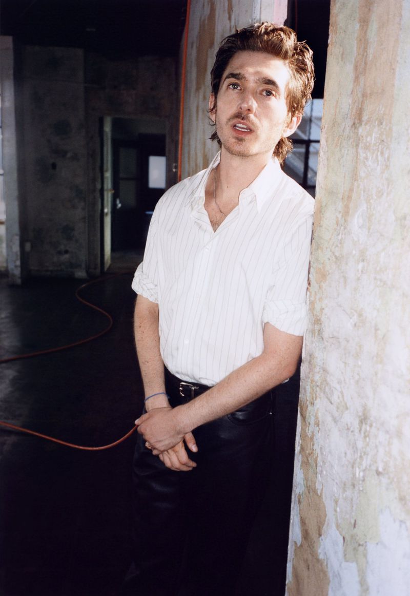 Austin Abrams leaning against a rough pillar, wearing a striped shirt and dark trousers for British GQ.