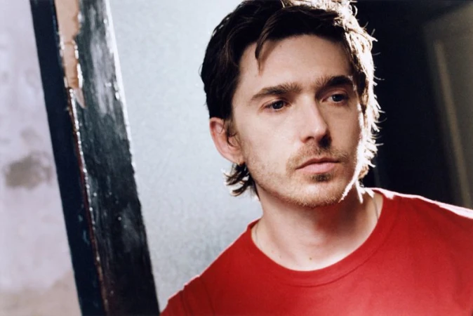 Austin Abrams wearing a red t-shirt in a portrait for British GQ, captured with natural lighting and moody tones.