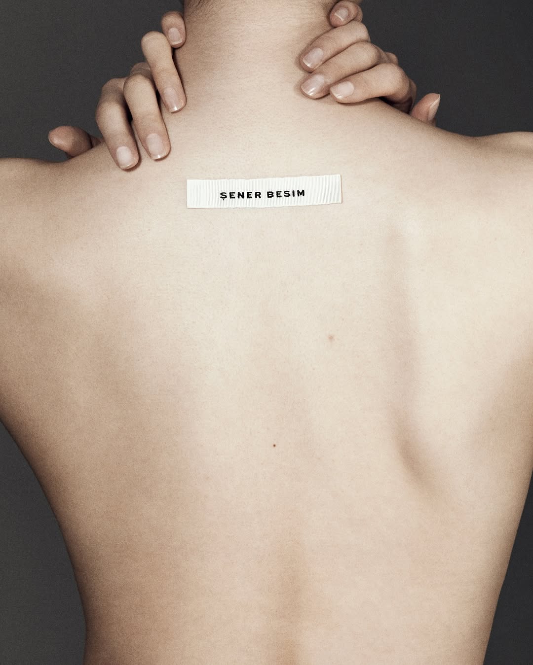 Back view of a person with fair skin and hands raised to the neck, featuring a 'ŞENER BESIM' label in the middle.