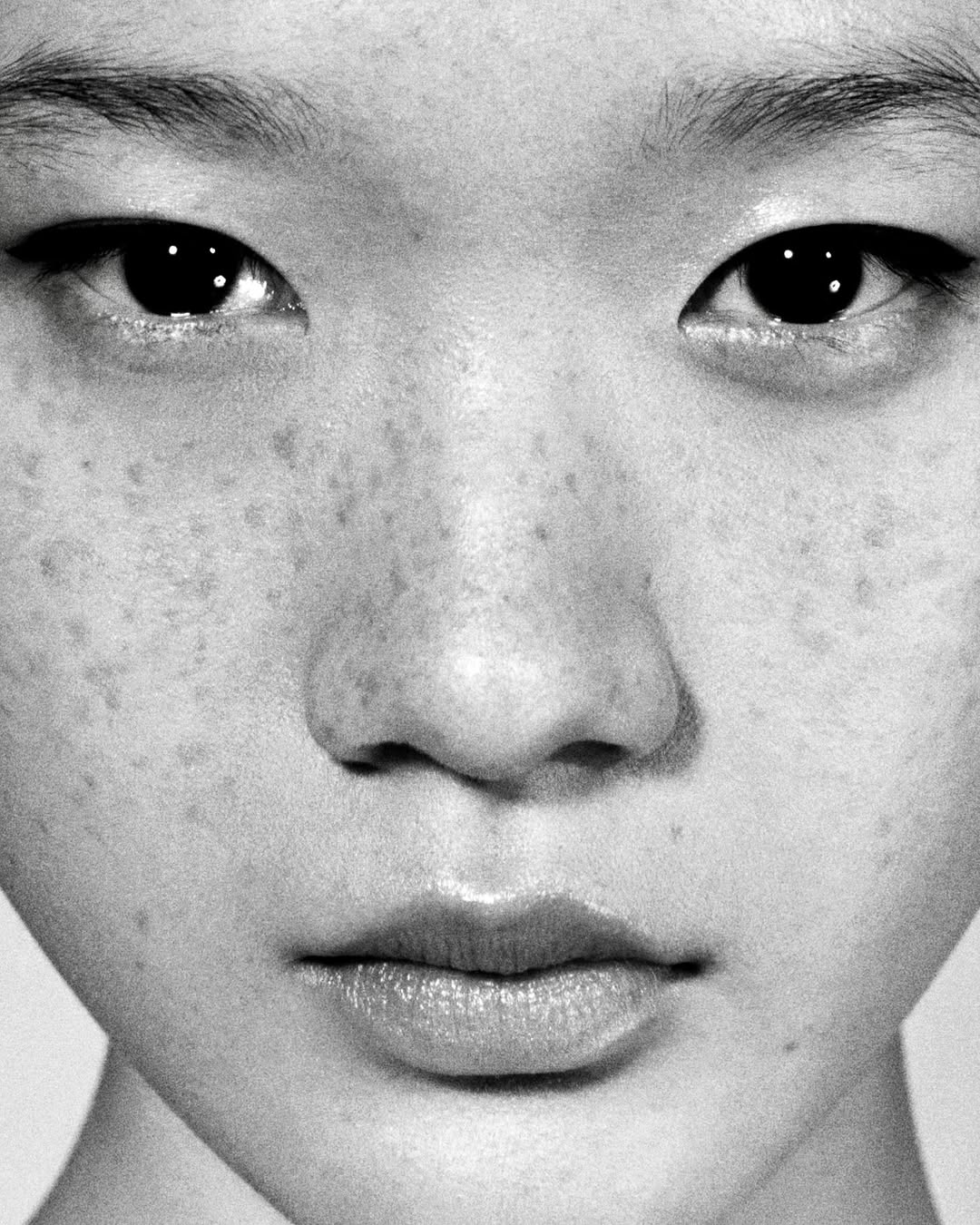 Close-up black and white portrait of a young Asian female with eyeliner and freckles, facing forward under natural light on a neutral background.
