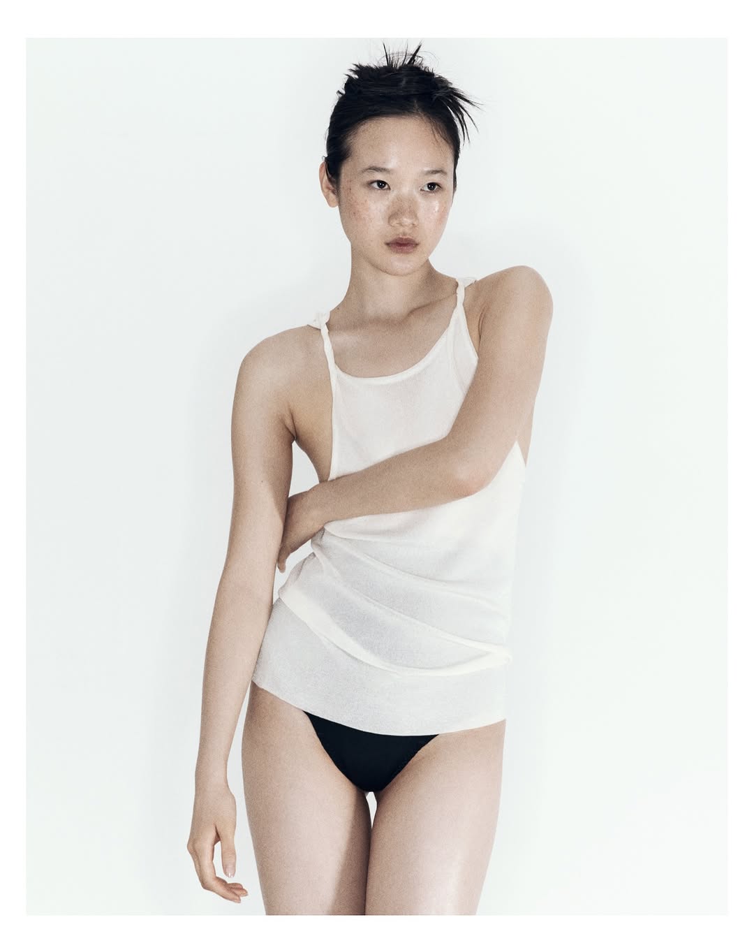 Portrait of a young woman with freckles posing in a white tank top and black underwear against a plain white background in a photo studio.