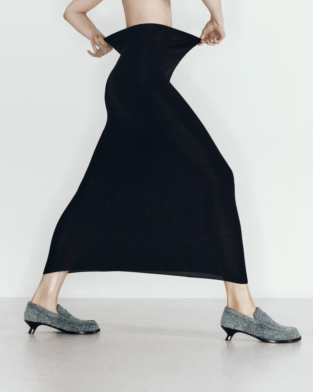 An adult female model poses in a long black skirt and silver shoes against a plain white background in a studio setting.