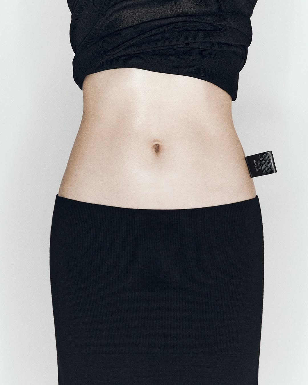 Close-up of a slim adult female torso wearing a black knit top and skirt, featuring a fabric label detail near the waist.