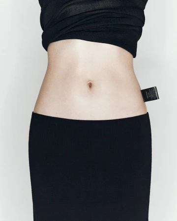 Close-up of a slim adult female torso wearing a black knit top and skirt, featuring a fabric label detail near the waist.