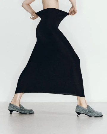 Model wearing a long black skirt holding the waistband and grey loafers against a white studio background.