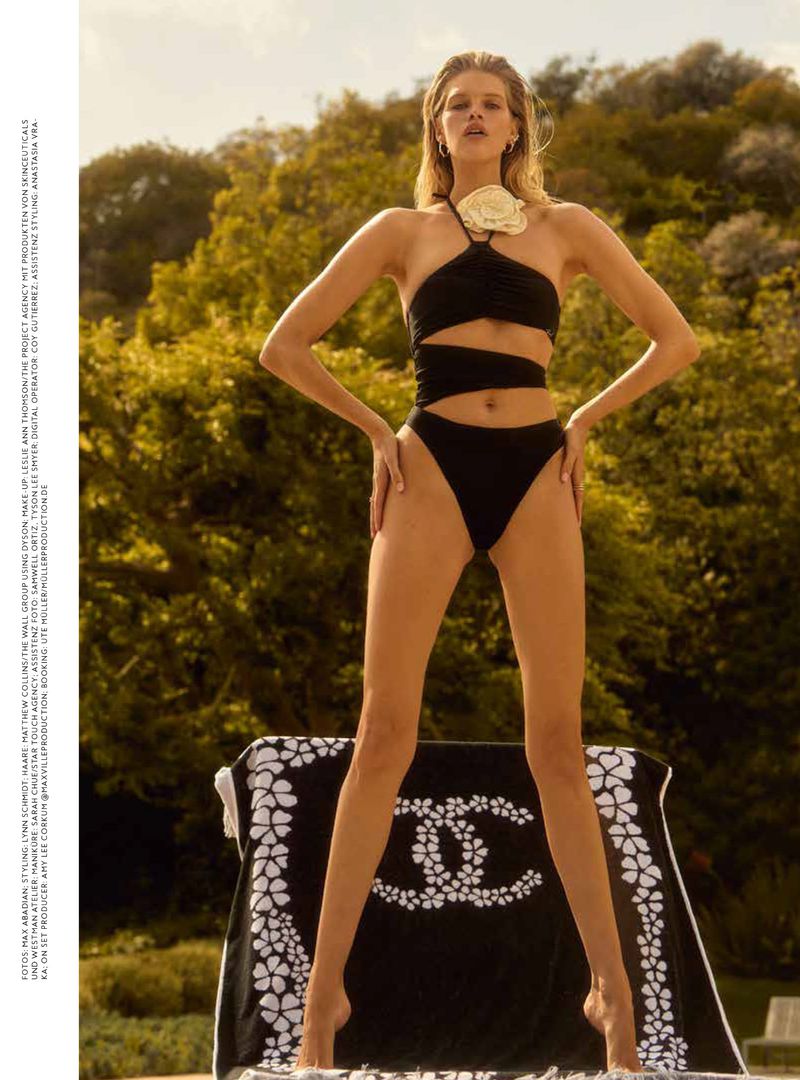 A slim adult woman poses in a black swimsuit with a flower accent on a patterned towel on a sunny day outdoors.