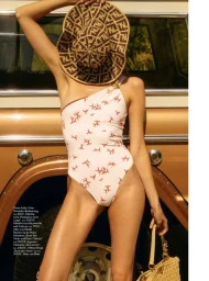 Model wearing a patterned one-piece swimsuit and holding a large straw hat in front of a vintage vehicle