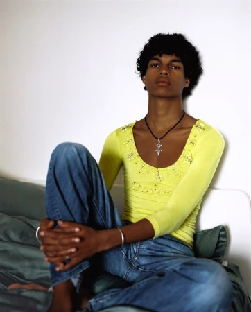 Young man with curly dark hair wearing a lime green scoop neck top and blue jeans seated against a white wall.