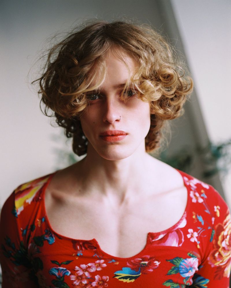 Portrait of a young man with curly blonde hair wearing a red floral print top against a plain studio wall