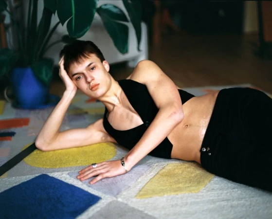 A young male model lying on a patterned carpet wearing a black crop top inside a residential interior.