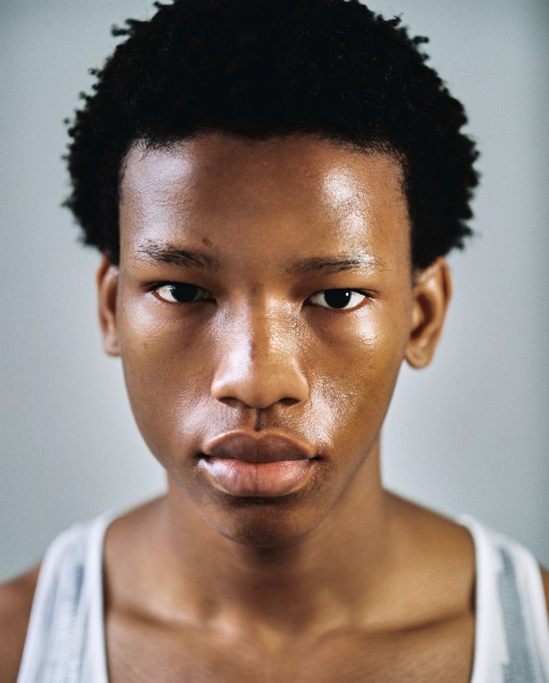 Close-up portrait of a young man with short dark curly hair wearing a white tank top against a neutral grey background.