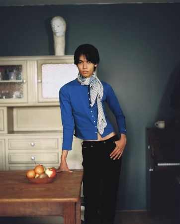 A man in a blue long-sleeve top and patterned scarf standing in a room with a wooden table, bowl of fruit, and a cabinet.