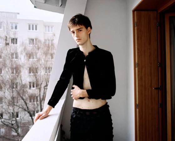 Young man in a black velvet jacket standing on a balcony overlooking an apartment building exterior.