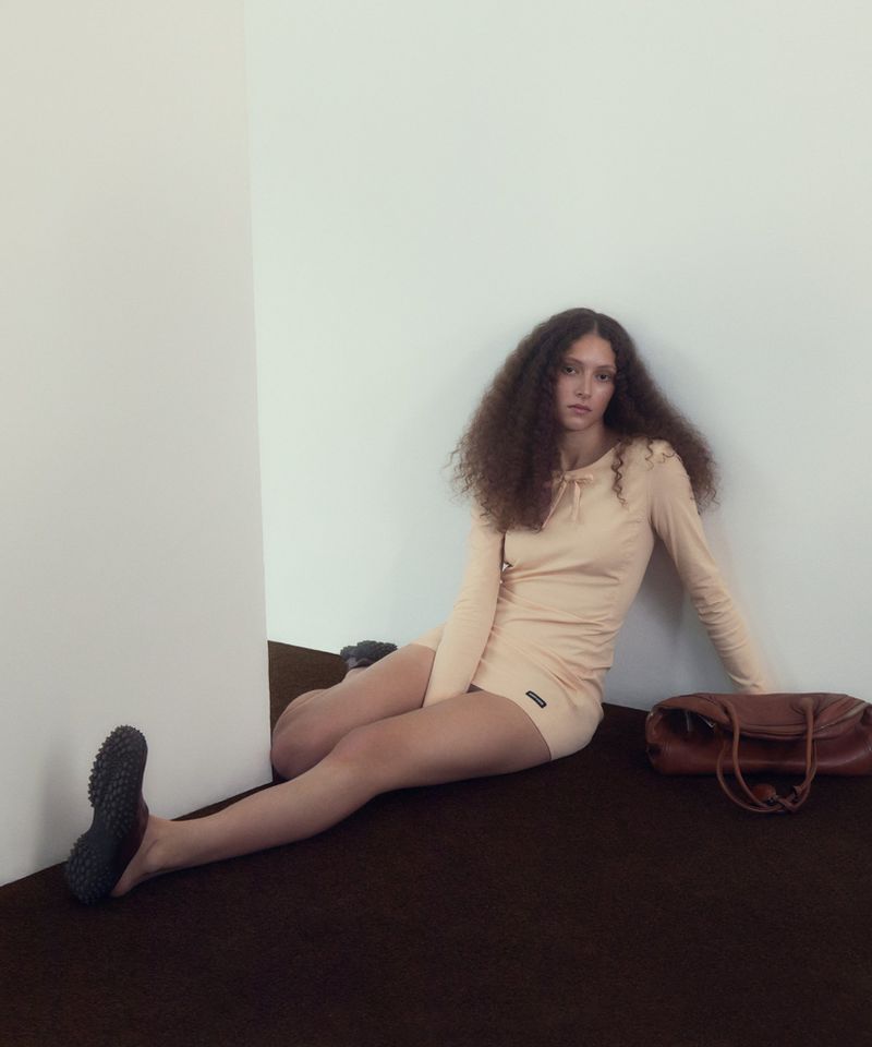 Model with long curly hair seated in a cream dress against a white wall next to a brown leather handbag.