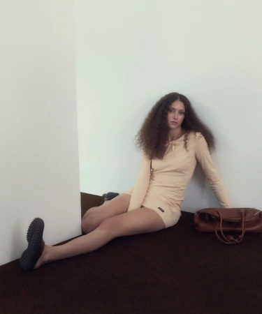Model with long curly hair seated in a cream dress against a white wall next to a brown leather handbag.