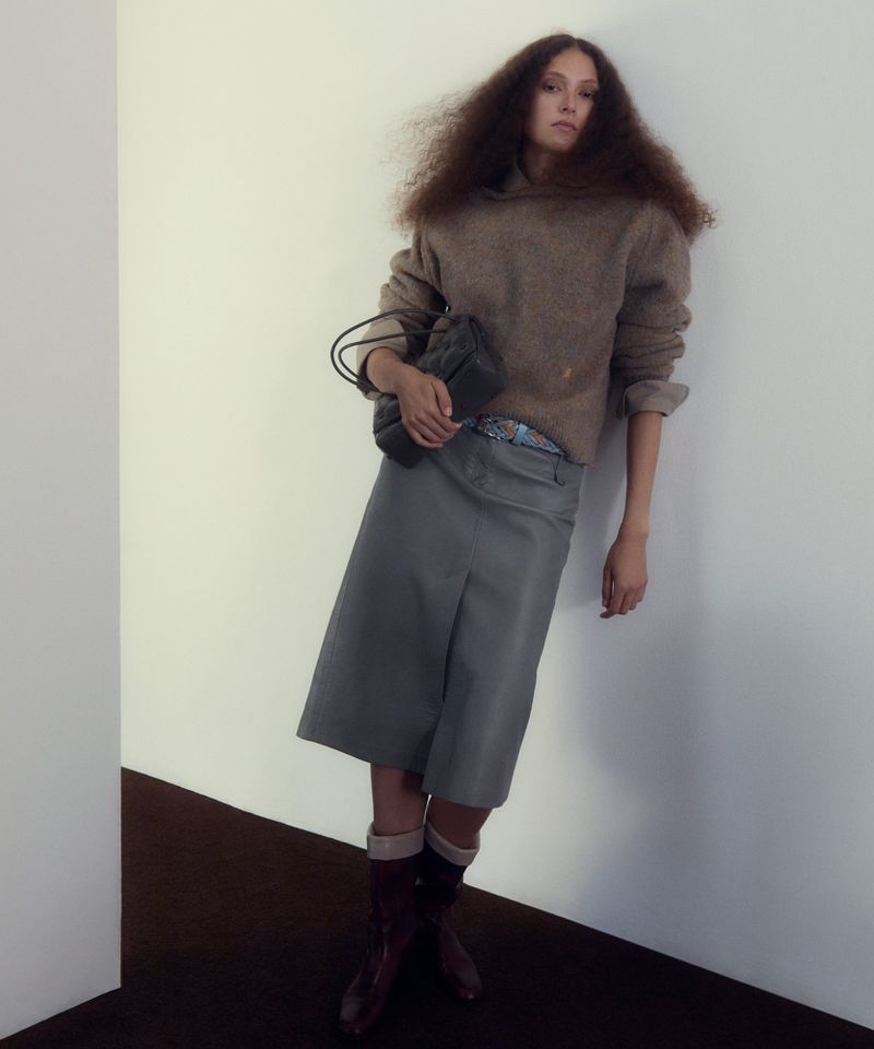Model with voluminous dark hair leaning against a plain white wall, wearing a brown sweater, grey skirt, and holding a dark handbag.