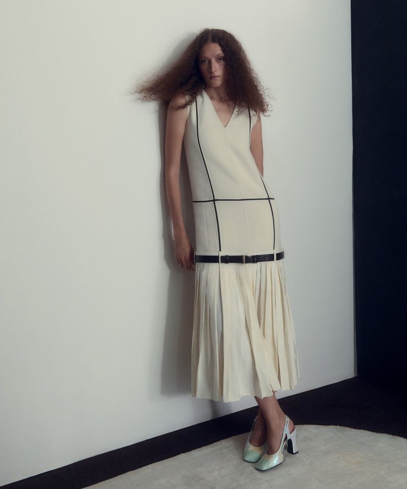 Model in a cream sleeveless midi dress with dark piping leaning against a white wall in a studio setting