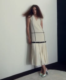Model in a cream sleeveless midi dress with dark piping leaning against a white wall in a studio setting
