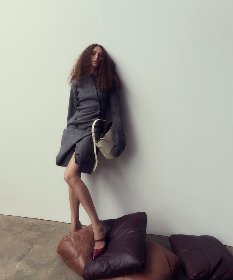 Model with long curly hair wearing a grey structured dress standing on leather cushions against a white background