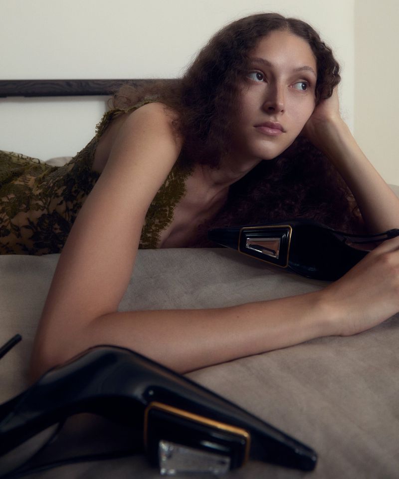 Model lying on a bed wearing a green lace dress with black pointed heels in the foreground, photographed by Daisy Walker.