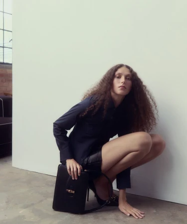 Model with long curly hair crouching in a dark shirt and black bag against a white wall