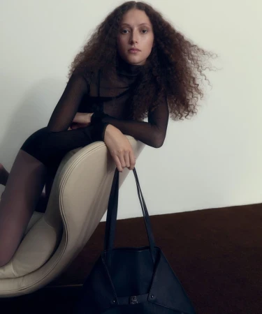 Model with long curly hair leaning over a curved chair holding a dark tote bag against a neutral background.