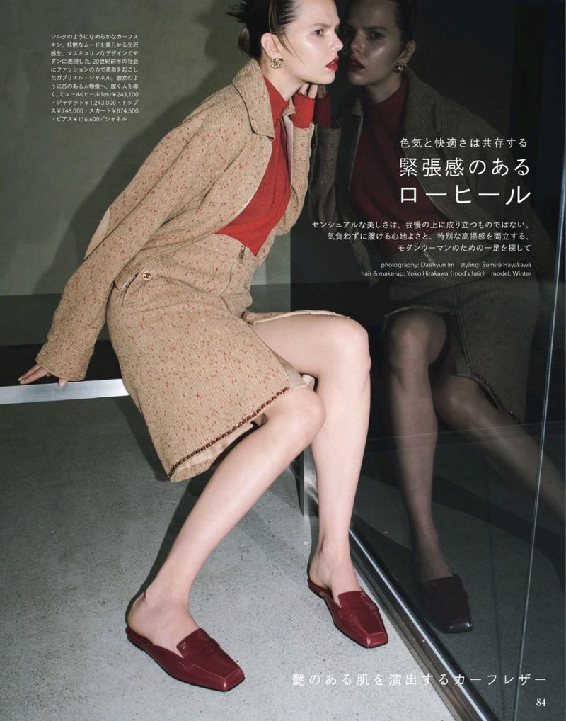 Model seated wearing a tweed jacket skirt suit with a red turtleneck and red leather mules in a reflective interior space