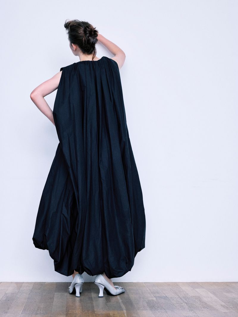 Model wearing a voluminous black dress and silver shoes against a plain white wall, captured from behind.