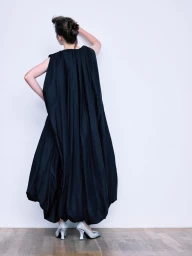 Model wearing a voluminous black dress and silver shoes against a plain white wall, captured from behind.